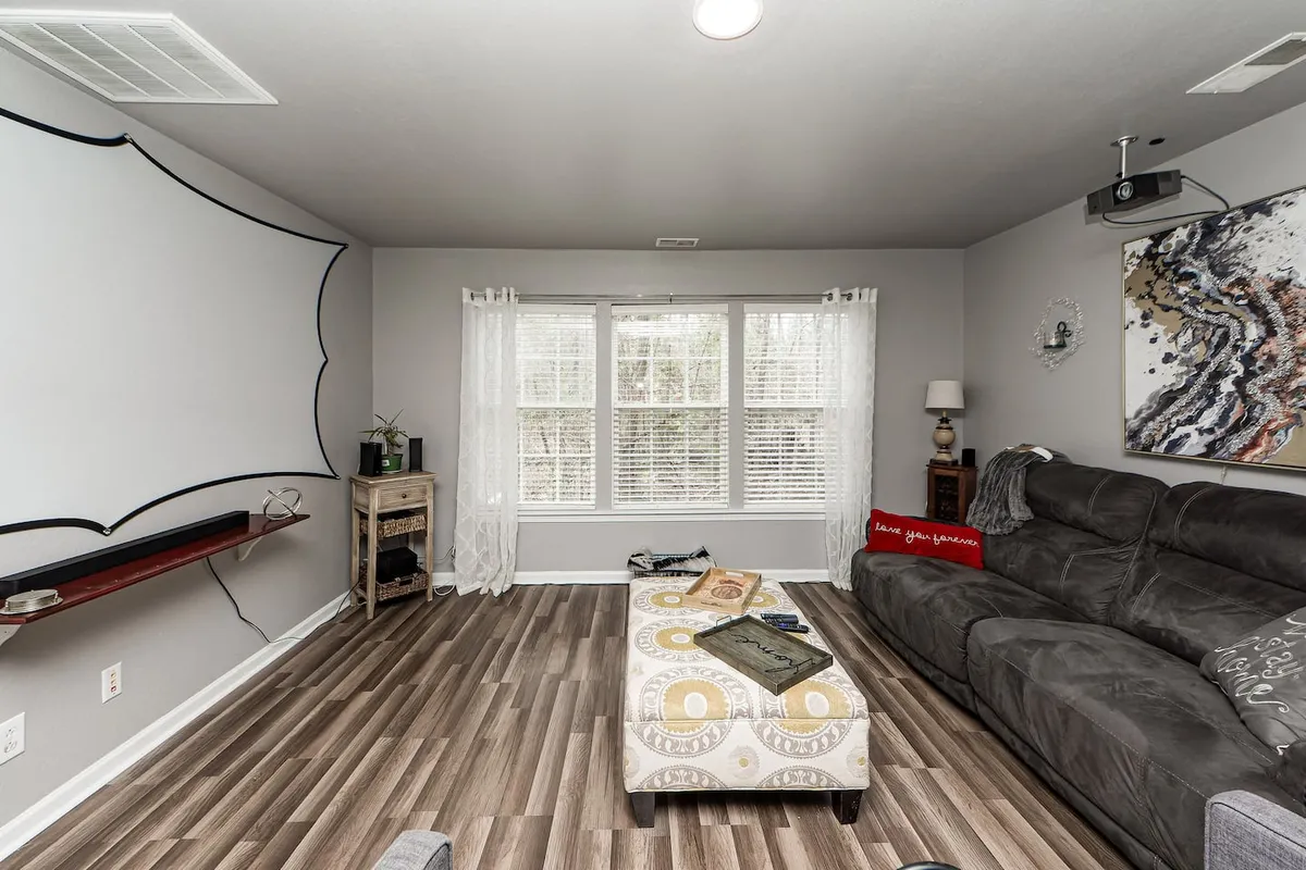 A cozy living room featuring a large window filling the space with natural light, a theater-style screen mounted on the wall, a comfortable sectional couch, and a decorative coffee table. The room exudes relaxation and is ideal for movie nights or gathering with friends.