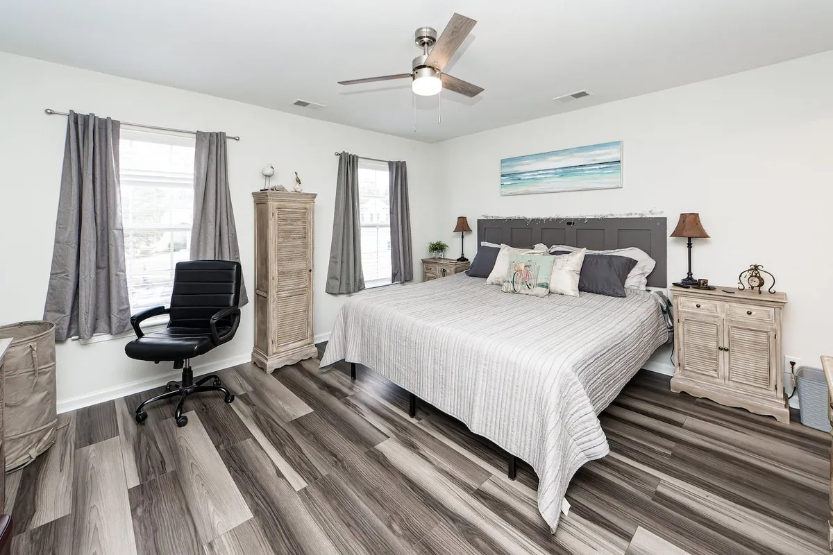 A serene bedroom with a neatly made bed, cozy nightstands, tasteful artwork, a window allowing in soft natural light, and a comfortable chair placed beside the window. The overall palette is calming and the room looks invitingly uncluttered, suggesting space for relaxation and reading.