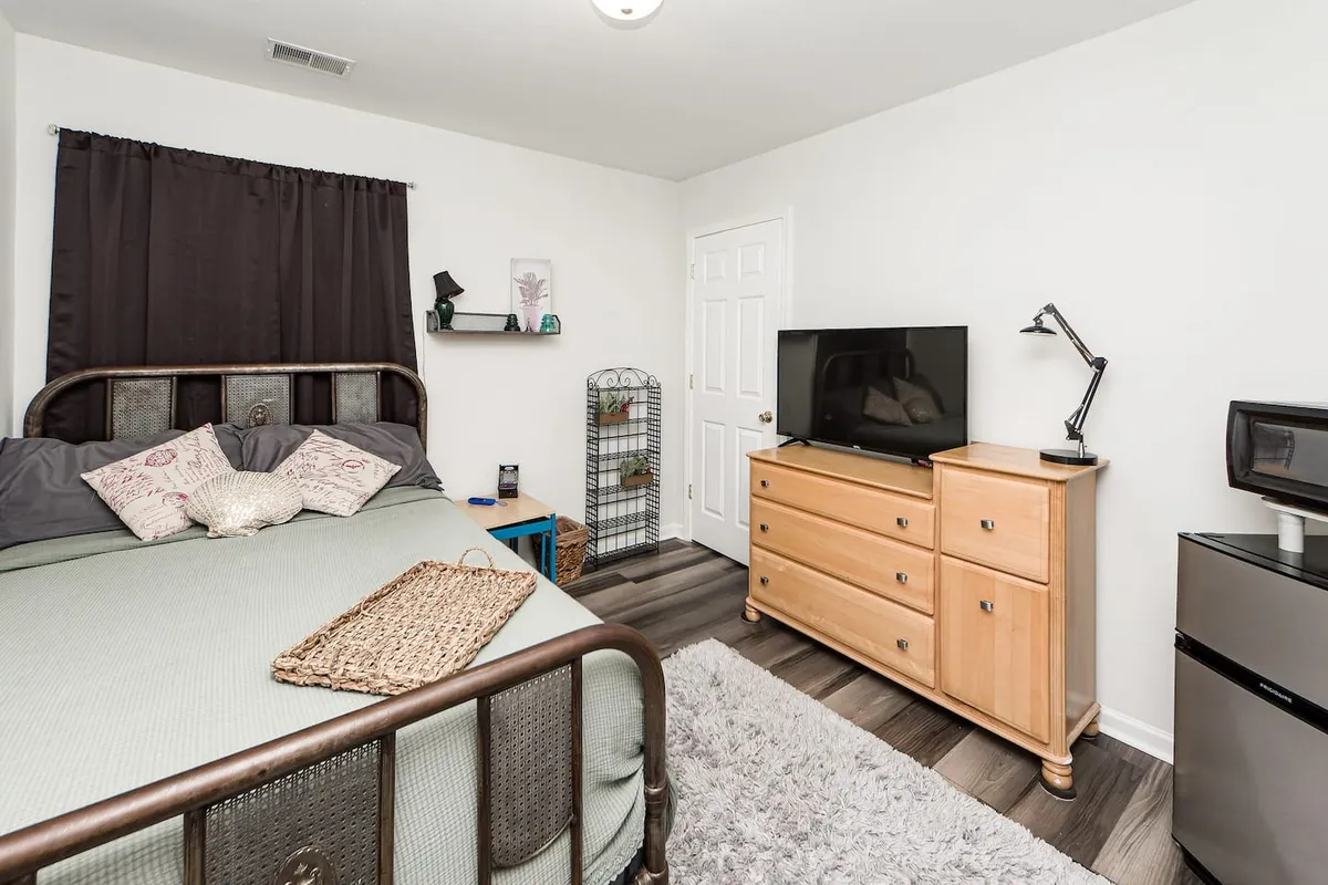 This bedroom features a cozy bed with decorative pillows, a flat-screen TV mounted above a wooden dresser, and shelves with decorative items. The atmosphere is intimate and comfortable, perfect for unwinding with personal streaming options or a quiet movie night.