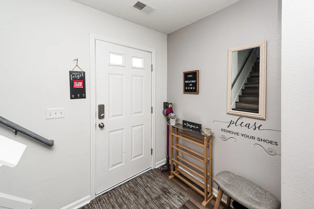 This image captures the home's front door area, complete with a welcome sign, shoe removal instructions, and a staircase visible in the background. The entryway is brightly lit and inviting, giving a sense of transitioning from the outside world into a cozy, welcoming space.