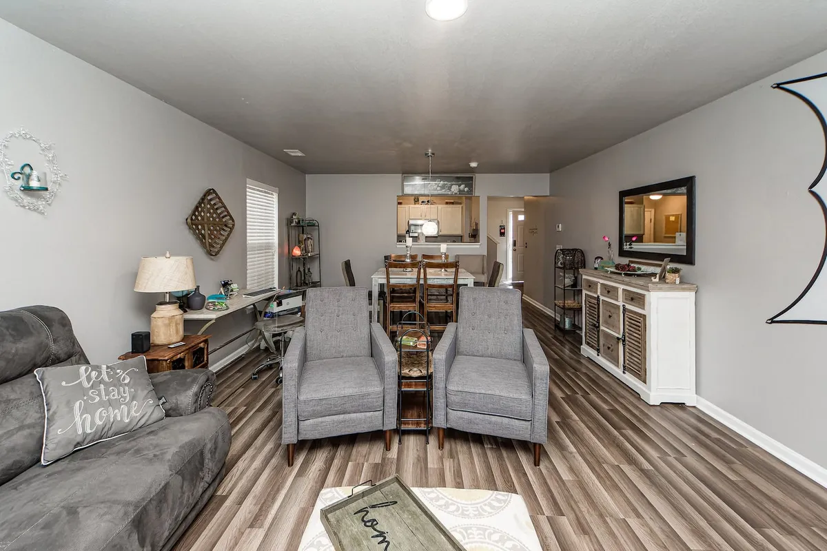A stylish lounge area with a comfortable gray sofa and two armchairs, a dining table visible in the background, decorative wall art, and a lamp casting a warm glow. The setting is designed for relaxation, socializing, and elegant gatherings.