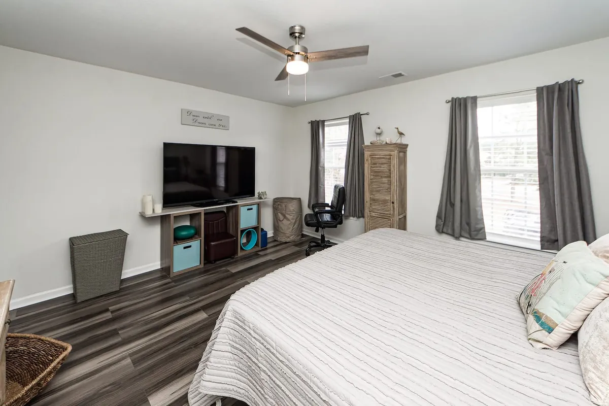 A serene bedroom with a cozy bed, a TV stand for entertainment, a small desk and chair for personal work or study, and a window allowing gentle natural light to fill the space. The decor is calm and contemporary, creating a personal retreat within the home.