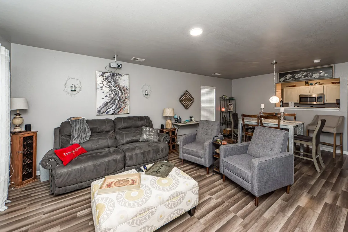 A warmly lit living space with a gray sectional sofa, a decorative ottoman, and a dining table with chairs in the background. Artistic wall décor and cozy accents make the room feel welcoming and perfect for a range of activities from entertaining guests to relaxing alone.